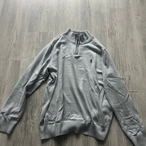 Ljusgrå half zip tröja Polo Ralph Lauren - Snygg ljusgrå långärmad tröja från Polo Ralph Lauren i storlek S. Tröjan har en stilren half zip-dragkedja vid halsen och klassisk broderad logga på bröstet. Mjuk bomullskvalitet och ribbade muddar vid ärmslut och nederkant.