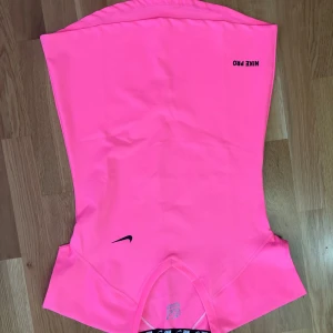 Nike Pro rosa träningströja - Nike Pro träningströja i en riktigt skarp rosa färg. Modellen är ärmlös och har en tight passform som sitter snyggt mot kroppen. Materialet är Dri-Fit med 92% bomull och 8% lycra, perfekt för intensiva träningspass. Diskret Nike-logga framtill och tryckt Nike Pro-text på axeln.