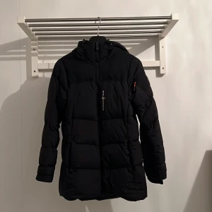Svart pufferjacka från Sail Racing - Svart pufferjacka från Sail Racing med huva och snygga detaljer som logopatch på ärmen och dragkedja framtill. Jackan har en clean look, är långärmad och har en orange zip-pull på ärmfickan. Perfekt för kalla dagar och har en schysst passform. Det finns även en ficka där tvättrådslappen sticker utifrån!