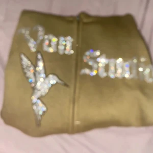 Olivgrön Vonstudio hoodie med strass - Olivgrön hoodie från Vonstudio i storlek M med strassdetaljer på bröstet, där det står 'Von Studio' och en glittrig kolibri. Tröjan har huva med snörning, hel dragkedja och mjukt material. Perfekt för dig som gillar statement-plagg och snygga detaljer.💚🫒