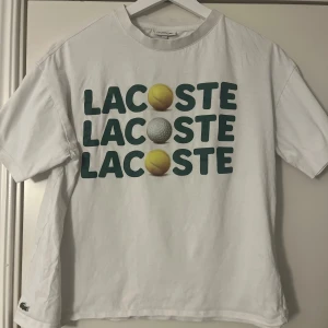 Vit Lacoste t-shirt med tryck - Vit t-shirt från Lacoste med tennis tema. Mycket sparsamt använd, typ 2 gånger. Inköpt för mindre än ett år sen, så rätt ny modell. Relaxed fit med korta ärmar och klassisk rund hals. 