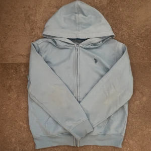 Ljusblå hoodie från U.S. Polo Assn. - Ljusblå hoodie från U.S. Polo Assn. med dragkedja framtill och broderad logga på bröstet. Tröjan har huva, långa ärmar och ribbade muddar. Perfekt för en chill och avslappnad stil. Pris kan diskuteras(: