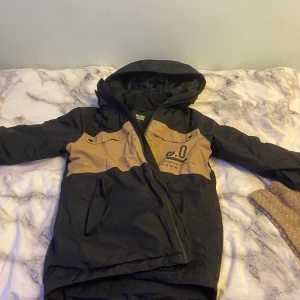 Svart och beige vindjacka Woxor 158 - Snygg vindjacka från Woxor i svart och beige med coolt tryck på bröstet och huva. Jackan har dragkedja framtill, fickor och detaljer som tryckknappar. Perfekt för dig som gillar streetstyle och vill ha en jacka som sticker ut.