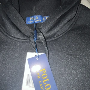 Svart hoodie från Polo Ralph Lauren - Svart hoodie från Polo Ralph Lauren med klassisk röd logobrodyr på bröstet. Tröjan har huva med snörning, stor magficka och långa ärmar. Materialet är mjukt och känns som bomull, perfekt för chill dagar. Snygg och enkel design med stilren look. Passar även M
