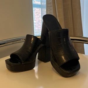Svarta plattformssandaler med klack - Säljer ett par svarta sandaler  med hög klack och plattform. Skorna har öppen tå och är gjorda i syntetmaterial med croco-präglad yta som ger en cool textur. Perfekt för dig som gillar chunky heels och statement skor.
