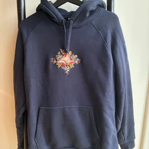 Mörkblå hoodie med broderi Les Deux - Snygg mörkblå hoodie från Les Deux med broderade blommor och text på bröstet. Tröjan har huva med snörning och en stor magficka. Perfekt för dig som gillar detaljer och vill ha en unik look. Storlek L men passar M. Nypris 1400