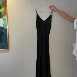 Svart långklänning från Melismee - Svart långklänning från Melismee Stockholm i 100% silke. Klänningen har smala axelband och v-ringad hals, vilket ger en stilren och elegant look. Silkesmaterialet gör den mjuk och följsam mot kroppen. Perfekt för dig som gillar minimalistisk och tidlös stil.