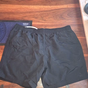 Svarta badshorts från SOC XL - Svarta badshorts från SOC i storlek XL. De har elastisk midja och är gjorda i lätt polyester med meshfoder för extra komfort. Perfekta för gymmet eller löprundan, med diskret logga på benet och praktiska sidofickor.