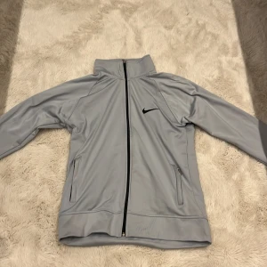 Ljusgrå vindjacka från Nike - Nike vindjacka i ljusgrått med svart broderad swoosh på bröstet. Jackan har hel dragkedja, höga muddar och två sidofickor med dragkedja. Materialet är slätt och syntetiskt, perfekt för träning eller chill. Långärmad och med sportig look.