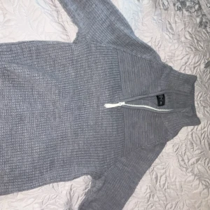 Grå stickad half zip-tröja Neomondo - Mysig grå stickad tröja från Neomondo med half zip och hög krage. Tröjan har ribbad struktur, långa ärmar och broderad logga på ena ärmen. Perfekt för lager-på-lager och chill dagar.