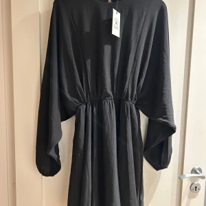 Svart långärmad klänning BikBok - Svart klänning från BikBok med markerad midja och vida, långa ärmar. Enkel och stilren design med rund halsringning och knapp i nacken. Tillverkad i ett mjukt, vävt material som ger ett snyggt fall. Perfekt för dig som gillar minimalistisk stil.