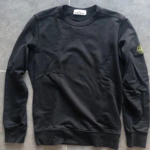 Svart sweatshirt från Stone Island - Svart sweatshirt från Stone Island med klassisk rund hals och långärmad passform. Tröjan har den ikoniska Stone Island-loggan på vänster ärm och är tillverkad i mjuk bomull. Perfekt för dig som gillar stilrena och snygga streetwear-plagg.