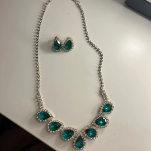 Set med halsband och örhängen med gröna stenar - Säljer ett glittrigt smyckeset med halsband och matchande örhängen. Båda har droppformade gröna stenar omgivna av små klara stenar i silverfärgad infattning. Perfekt för dig som vill sticka ut med lite extra bling.