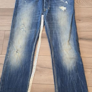Blå DNA dirtynewage jeans med slitningar - Säljer ett par blå jeans från DNA dirtynewage med coola slitningar och blekta detaljer på benen. Klassisk femficksmodell med raka ben och gul söm på bakfickorna. Jeansen har normal passform och är tillverkade i jeansmaterial.