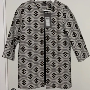 Mönstrad kavaj från Vero Moda - Säljer en snygg kavaj från Vero Moda i storlek 36. Jackan har ett grafiskt mönster i svart, vitt och guld, raka linjer.  Modellen är rak och har ingen krage, vilket ger en clean och modern look. Perfekt för dig som gillar att sticka ut.