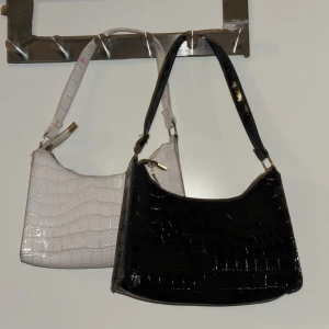 Svart och vit axelväska med croco-mönster - Två stilrena axelväskor i svart och vit med glansig yta och croco-mönster. Väskorna har en enkel, rektangulär form och dragkedja upptill. Perfekt för dig som gillar trendiga accessoarer med lite edge. Säljer båda för 20 kr