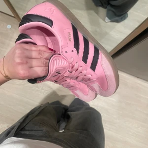 Adidas samba rosa sneakers - Säljer ett par Adidas Messi sneakers i en snygg ljusrosa färg med svarta ränder och detaljer. Skorna har klassisk snörning, platt sula och rund tå. Ovandelen är i skinn och mocka, med gummisula. Perfekt för dig som vill sticka ut med färg på fötterna.