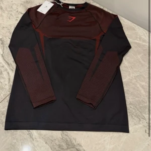 Onyx V5 red - Svart långärmad träningströja från Gymshark med mörkröda detaljer på axlar och ärmar. Tröjan har ett sportigt snitt och är tillverkad i ett lätt syntetmaterial som andas. Perfekt för gymmet eller löprundan, med Gymshark-logga på bröstet.