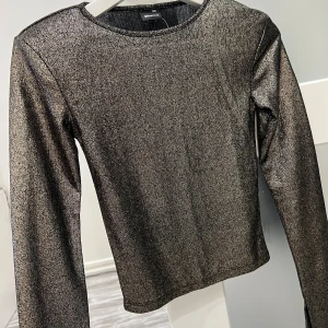 Glittrig långärmad topp Gina Tricot - Säljer en glittrig långärmad topp från Gina Tricot i storlek XXS/XS.Toppen har en rund halsringning och är i en mörk silverfärg med skimrande effekt. Materialet är syntet och ger en cool metallic look som verkligen sticker ut.