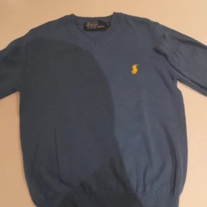 Blå stickad tröja Polo Ralph Lauren - Säljer en blå stickad tröja från Polo Ralph Lauren med gul broderad logga på bröstet. Tröjan har v-ringning, ribbade muddar och lång ärm. Perfekt för dig som gillar klassisk och stilren look.