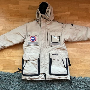Beige Canada Goose dunjacka med huva - Säljer en beige dunjacka från Canada Goose med svart foder och detaljer. Jackan har flera stora fickor, patchar på bröstet och svarta förstärkningar på armbågarna. Justerbar huva med snörning och dragkedja framtill. Perfekt för kalla dagar.Canada Goose Snow Mantra, Super härlig och varm jacka.  Storlek S/P. Det är inte storlek small utan större. Skulle säga mer M-L.  Päls med följer.  Inga skador i övrigt, se sista bilderna dock. Snoddarna är lite uttöjda men funkar. Jackan är välanvänd så tecke
