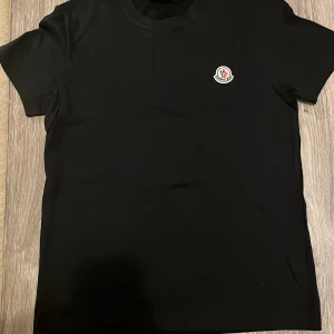 Svart Moncler t-shirt med logga - Svart t-shirt från Moncler med klassisk rund hals och korta ärmar. Diskret Moncler-logga på bröstet ger en clean och exklusiv vibe. Tillverkad i mjuk bomull för maximal komfort. Perfekt för dig som gillar stilrena och lyxiga plagg.