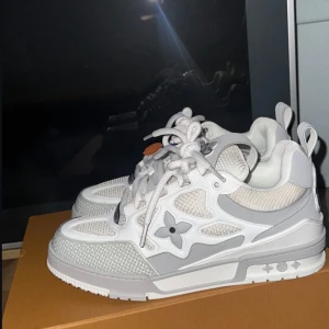 Louis Vuitton vita sneakers med grå detaljer - Snygga Louis Vuitton sneakers i vitt och ljusgrått med ikonisk blomdetalj på sidan. Skorna har rund tå, platt sula och snörning. Materialmix av skinn och mesh ger en modern look. Perfekta för dig som gillar exklusiva märken och stilren design.