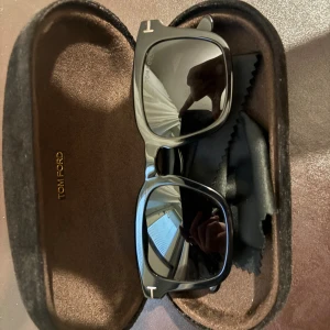 Solglasögon Tom Ford - Snygga svarta solglasögon från Tom Ford med rektangulär form och diskret T-detalj i metall på bågen. Glasögonen är tillverkade i Italien och har en stilren, modern look. Kommer med originalfodral och putsduk.