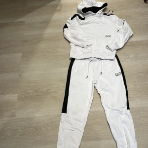 Vit hoodie/byxa från EA7 med svarta detaljer - Säljer en snygg vit hoodie/byxa set från EA7 med svarta detaljer på axlar/ben och huva. Tröjan har huva med dragsko och EA7-logga på bröstet. Perfekt för dig som gillar sportig och clean stil. Finns 2 små fläckar en på byxa en på tröja!!