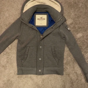 Grå hoodiejacka från Hollister S - Mysig grå hoodiejacka från Hollister med blå quiltad insida och fluffigt foder i huvan. Jackan har dragkedja, knappar och två stora fickor framtill. Perfekt för dig som gillar chill och streetstyle.