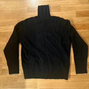 Svart polotröja med lång ärm - Svart polotröja med hög krage och långärmad design. Tröjan är enkel och stilren, perfekt för lager-på-lager och har en mjuk känsla. Passformen är normal och materialet känns som bomull eller syntet. Snygg att matcha med jeans eller kjol.