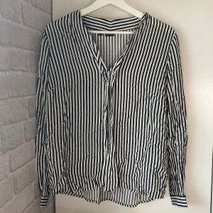 Blus - Snygg tunnare långärmad blus från Bodyflirt i svart och vitt med vertikala ränder. Blusen har v-ringning och knytband framtill för extra stilpoäng. Perfekt för dig som gillar en clean och trendig look.