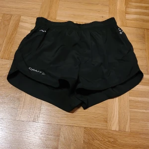 Svarta träningsshorts från Craft - Svarta sportiga shorts från Craft med elastisk midja och diskret logga framtill. Lätta och luftiga med kort passform, perfekta för löpning eller gym. Materialet är syntet och designen är enkel utan extra detaljer. Snygga och funktionella för aktiva dagar.