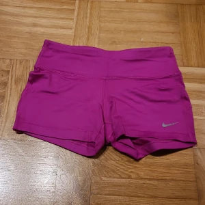 Nike Dri-Fit rosa träningsshorts XS - Snygga och sportiga rosa träningsshorts från Nike i Dri-Fit material. Modellen är högmidjad och har en liten ficka med dragkedja baktill. Perfekta för gymmet eller löpning, med en tight passform och diskret Nike-logga framtill.