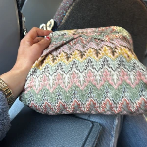 Mönstrad kuvertväska i pastell - Kuvertväska med färgstarkt zigzag-mönster i pastellfärger som rosa, grön, gul och vit. Väskan har mjuk form och är tillverkad i ett tygmaterial med broderad känsla. Perfekt för att addera lite färg till din outfit.
