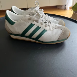 Adidas Country sneakers vit/grön - Säljer ett par klassiska Adidas Country sneakers i vitt skinn med gröna ränder och detaljer. Skorna har snörning, mockadetaljer vid tån och häl samt gummisula med fiskbensmönster. Perfekta för dig som gillar retrostil och sportig vibe.