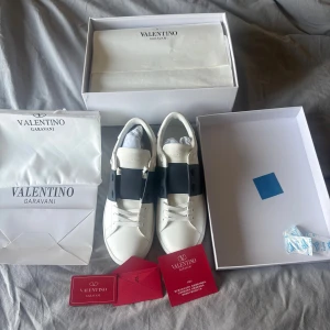 Valentino Garavani Open sneakers mörkblå - Helt oanvända snygga Valentino Garavani Open sneakers i vitt skinn med svart band över snörningen. Klassisk rund tå, platt sula och diskret guldlogga på plösen. Kommer med originalkartong, dustbag och äkthetskort. Perfekta för dig som gillar lyxiga och stilrena sneakers. 
