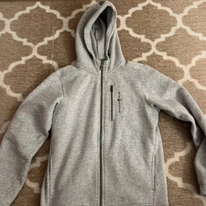 Ljusgrå hoodiejacka Sail Racing - Ljusgrå hoodiejacka från Sail Racing med hel dragkedja och extra zipficka på bröstet. Jackan har mjukt fleecefoder, lång ärm och normal passform. Perfekt för chill dagar och har snyggt broderad logga på framsidan.