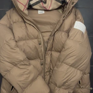 Beige pufferjacka från Burberry - Säljer en beige pufferjacka från Burberry med stor patch på ärmen där det står 'Burberry London England'. Jackan har dragkedja och tryckknappar framtill, två sidofickor med dragkedja och en mysig, oversized passform. Materialet är polyester och jackan har långa ärmar.