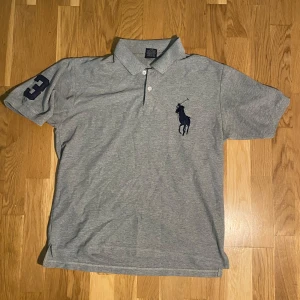 Polo Ralph Lauren piké - Snygg grå pikétröja från Polo Sport i 100% bomull. Klassisk krage med två vita knappar, stor mörkblå polospelare broderad på bröstet och en mörkblå siffra 3 på ärmen. Perfekt för en chill och sportig look.