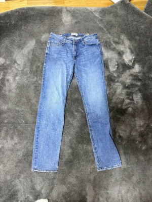 Blå jeans från Jack & Jones - Snygga blå jeans från Jack & Jones med klassisk femficksdesign och ljus tvätt. Modellen har raka ben och normal passform, med bruna sömmar och läderpatch bak i midjan. Perfekt för dig som gillar stilrena och tidlösa jeans.