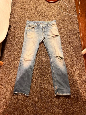 Tommy Hilfiger Ryder Zip jeans ljusblå - Ljusblå jeans från Tommy Hilfiger Denim, modell Ryder Zip. De har slitna detaljer och hål vid knäna för en cool, vintage vibe. Klassisk femficksdesign, raka ben och dragkedja i gylfen. Materialet är jeans i bomull.