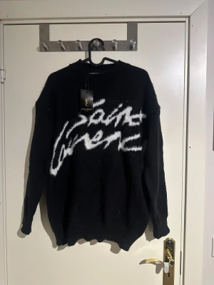 Svart stickad tröja Yves Saint Laurent - Svart stickad tröja från Yves Saint Laurent med vit, fluffig logotyp i graffiti-stil över bröstet. Tröjan har rund hals, ribbade muddar och lång ärm. Perfekt för dig som gillar statement-plagg och exklusiv streetwear.