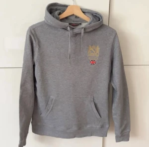 Grå hoodie från Morris - Grå hoodie från Morris London med broderad guldfärgad logga och brittisk flagga på bröstet. Tröjan har huva med snörning och en stor magficka. Perfekt för dig som gillar klassisk och stilren streetwear.