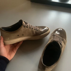 Bruna lanvins  - Säljer ett par bruna sneakers med snygg kombination av mocka och lackad tå. Skorna har snörning och platt sula, vilket ger en clean och stilren look. Perfekta för dig som gillar en neutral färgskala med en twist. Passar till det mesta i garderoben. Det ända felet med dem är att det är en liten skada på tån men det är bild på det och sen är inte större än en halv cm. Dem är 42 men passar 43 nästan bättre skulle jag säga. Byten är ett stort intresse!