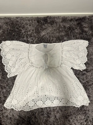 Vit broderad blus från Lindex - Superfin vit blus från Lindex med broderade detaljer och vida, korta ärmar. Blusen har knytning framtill och vackert hålmönster längs kanterna och ärmarna. Perfekt för varma dagar och ger en romantisk känsla till din outfit.