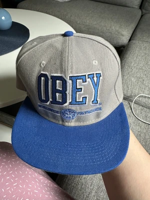 Grå och blå snapback från Obey - Snygg snapback-keps från Obey i grått med blå skärm och broderade detaljer. Framsidan har stor blå OBEY-text och coolt emblem, baksidan har en blå stjärna med krona. Materialet är 80% akryl och 20% ull. Perfekt för streetwear-stilen.