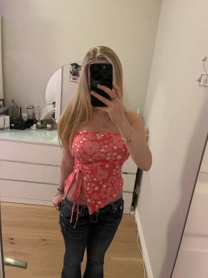 Rosa blommig tubtopp med snörning - Säljer en rosa tubtopp med blommigt mönster och vita detaljer. Toppen har snörning på sidan och en asymmetrisk spetsig nederkant. Materialet är mjukt och stretchigt, perfekt för sommarens alla häng. Sitter tight och är axelbandslös för en snygg look. Knappt använd därför säljer jag, det står ingen storlek men jag skulle gissa på xs/s. 