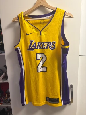 Gul Lakers Ball #2 baskettröja Nike S - Säljer en officiell Los Angeles Lakers baskettröja med Ball och nummer 2 på ryggen. Tröjan är gul med lila och vita detaljer, ärmlös och tillverkad i Dri-Fit material som andas. Lakers-logga och Nike Swoosh på framsidan. Perfekt för dig som älskar basket och streetwear. Tröjan är äkta. Lonzo ball