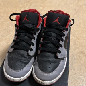 Nike Air Jordan 1 Low svart/röd/grå - Nike Air Jordan 1 Low sneakers med svart och grå skinnöverdel, röda detaljer och röd Swoosh på sidan. Klassisk Jumpman-logga på tungan och Air Jordan-märke på hälen. Låg modell med svart snörning och vit/röd yttersula. Perfekt för dig som gillar streetwear och ikoniska sneakers. Använd mycket 